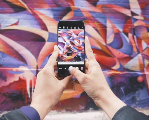 A pair of hands holding a mobile phone, taking a photograph of colourful graffiti